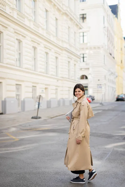 Happy woman in beige trench coat walking with laptop on street in vienna — Stock Photo