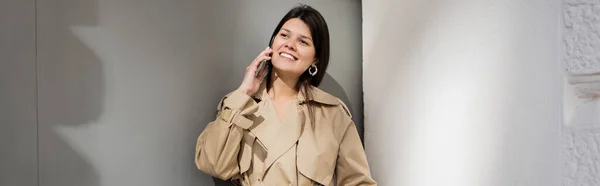 Happy woman in trench coat standing near wall and talking on smartphone, banner — Stock Photo