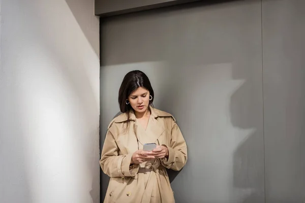 Brunette woman in trench coat texting on smartphone near wall — Stock Photo