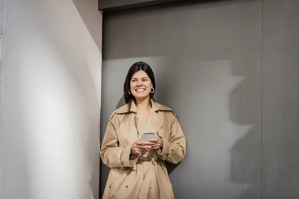 Happy woman in trench coat texting on smartphone near wall — Stock Photo