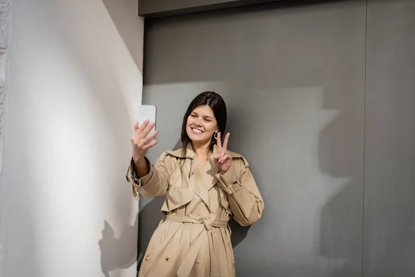 Happy woman in trench coat taking selfie and showing peace sign near wall — Stock Photo