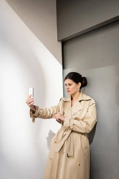 Brunette woman in trench coat taking selfie on smartphone near wall — Stock Photo