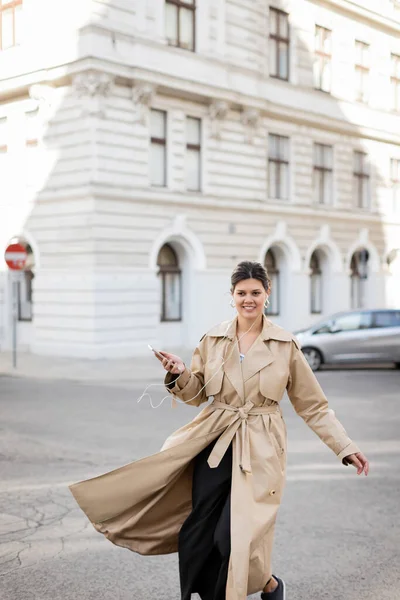 Cheerful woman in wired earphones holding mobile phone and listening music on street in vienna — Stock Photo