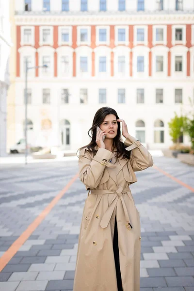 Morena mujer en abrigo beige hablando en smartphone en la calle de vienna - foto de stock