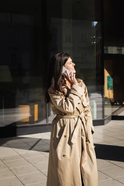 Cheerful woman in beige coat talking on smartphone on street of vienna — Stock Photo