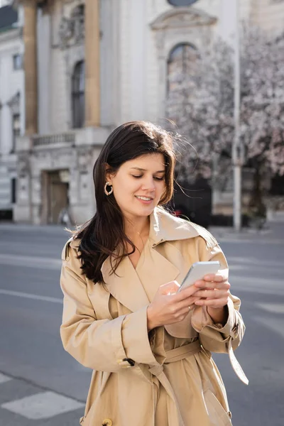 Donna soddisfatta in cappotto elegante utilizzando smartphone sulla strada europea — Foto stock