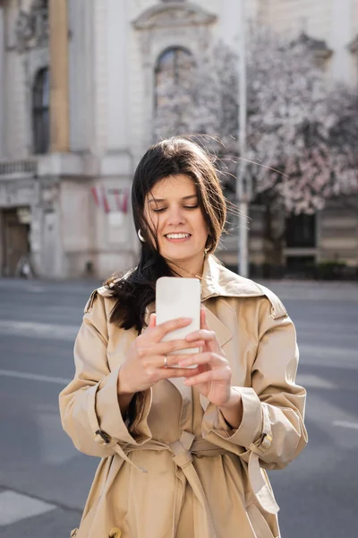 Mujer alegre en abrigo elegante usando teléfono inteligente en la calle europea - foto de stock
