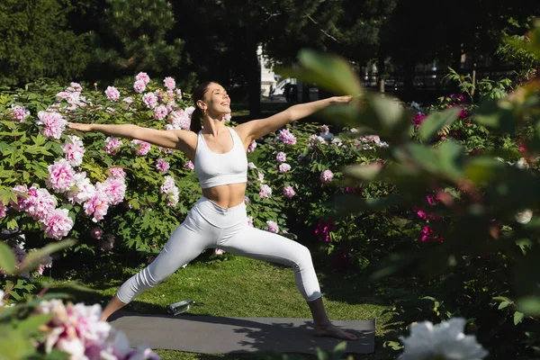 Happy sportive woman practicing warrior pose with outstretched hands in park — Stock Photo