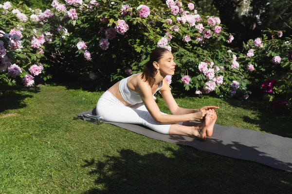 Slim woman practicing seated forward bend pose on yoga mat near blossoming bushes — Foto stock