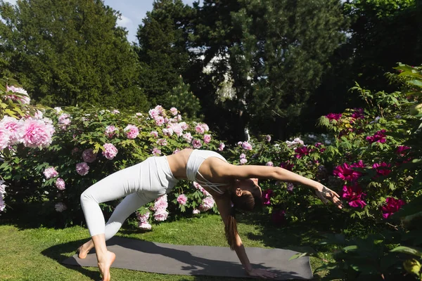Стройная женщина в белой спортивной одежде практикующая дикую позу йоги возле цветущих растений в парке — стоковое фото