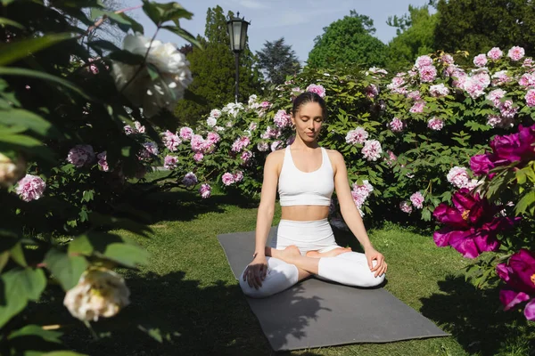 Woman with closed eyes meditating in lotus pose near blossoming plants in park — Stockfoto