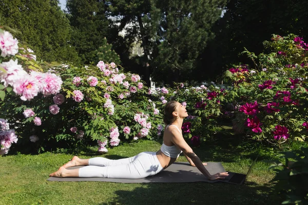 Vue latérale de la femme heureuse pratiquant le yoga en cobra pose sur la pelouse près d'un ordinateur portable — Photo de stock