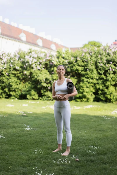 Barfüßige Frau in weißer Sportbekleidung steht mit Yogamatte auf grünem Rasen — Stockfoto