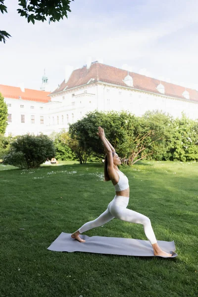 Side view of sportive woman practicing crescent lunge pose on yoga mat in city park - foto de stock