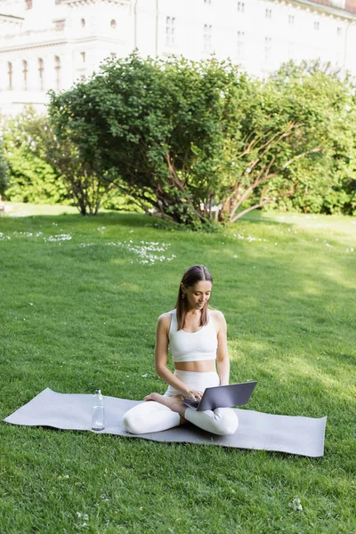 Woman using laptop while sitting in lotus pose on yoga mat and green lawn in park — Stock Photo