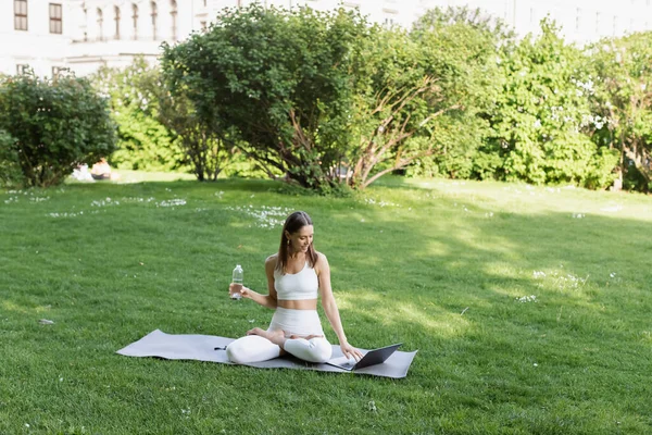 Woman in white sportswear sitting in lotus pose near laptop in park — Stock Photo