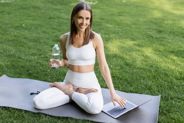 Joyful woman with sports bottle looking at camera while sitting in lotus pose near laptop — Stock Photo
