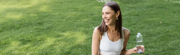 Mujer feliz en sujetador deportivo blanco sosteniendo botella deportiva y mirando al aire libre, pancarta - foto de stock