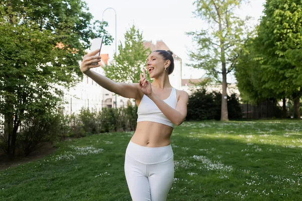 Cheerful sportswoman sticking out tongue and showing victory sign while taking selfie in park - foto de stock
