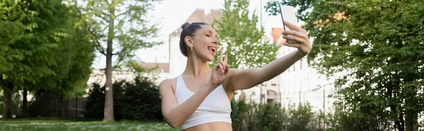 Sportive woman showing victory gesture and sticking out tongue while taking selfie in park, banner — Photo de stock
