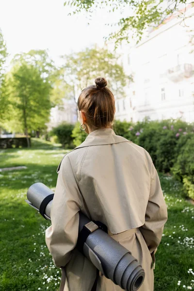 Back view of woman in trench coat holding fitness mat in city park - foto de stock