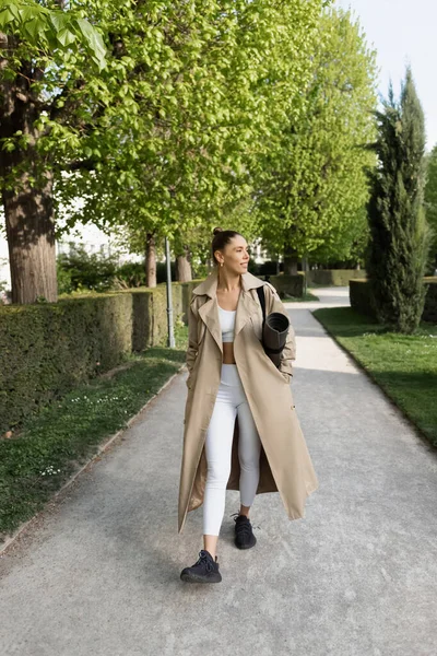 Full length of smiling sportswoman in trench coat walking with fitness mat in park — Stock Photo