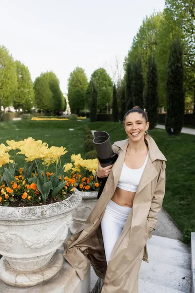 Femme gaie en vêtements de sport et trench coat tenant tapis de fitness près des vases du parc avec des fleurs — Photo de stock