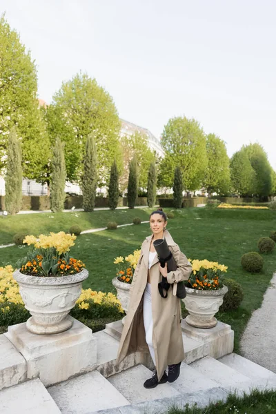 Sportswoman in trench coat holding fitness mat while walking on stairs near park vases with flowers — стоковое фото