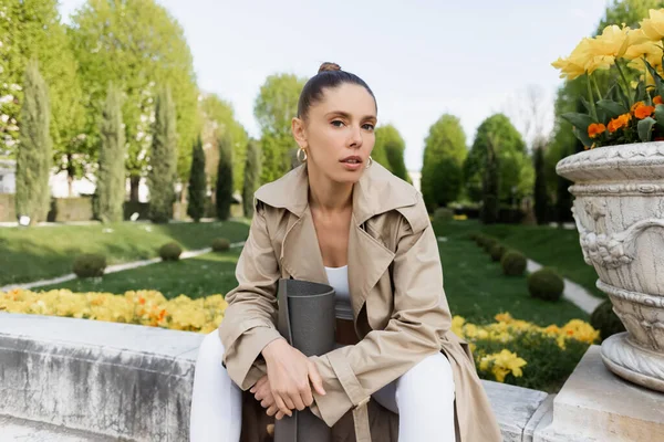 Femme en trench coat regardant la caméra tout en étant assis avec tapis de fitness près du vase de parc — Photo de stock