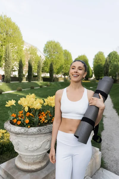 Joyful woman in sports bra holding fitness mat near park vase with blossoming flowers — Stock Photo