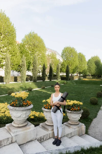 Full length of sportswoman holding fitness mat while standing on stairs near park vases — стоковое фото