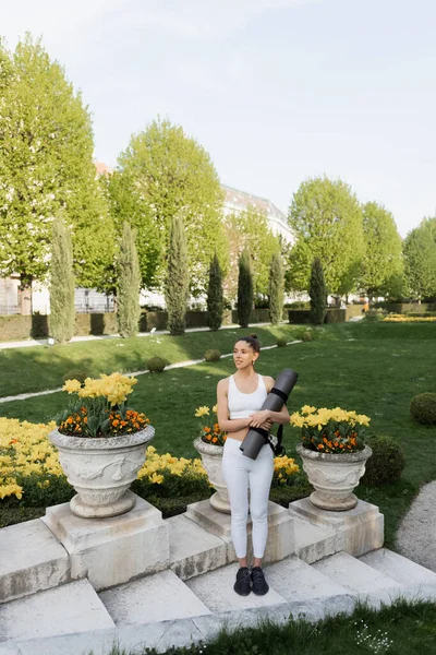 Comprimento total da mulher em sportswear branco de pé com tapete de fitness perto de vasos do parque com flores — Fotografia de Stock