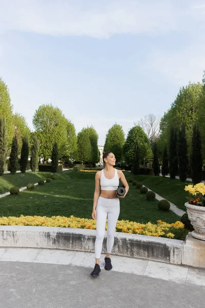 Женщина в белом в полный рост носит фитнес-коврик и смотрит вдаль в парке — стоковое фото