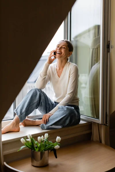 Cheerful barefoot woman in jeans sitting on windowsill near tulips and talking on mobile phone — Foto stock