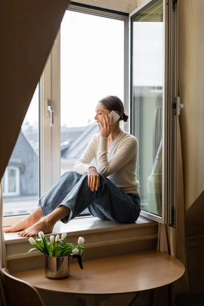 Barfüßige Frau in Jeans unterhält sich auf Fensterbank in der Nähe von Tulpen mit dem Handy — Stockfoto
