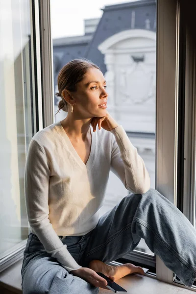 Verträumte Frau schaut weg, während sie zu Hause auf der Fensterbank sitzt — Stockfoto