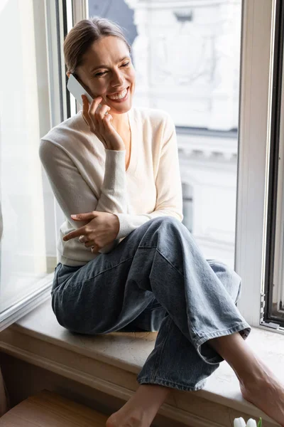 Joyful woman sitting on windowsill during conversation on mobile phone — Photo de stock