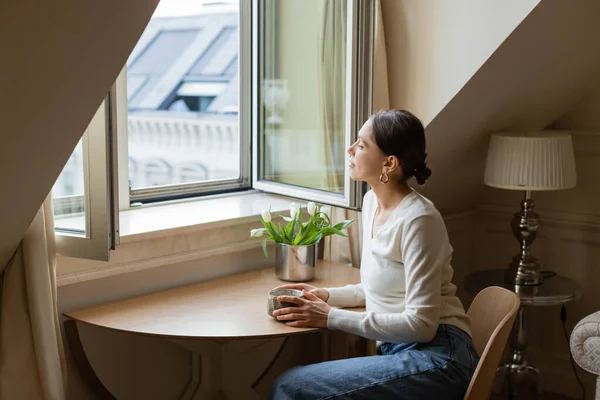 Mujer pensativa sentada con taza de arcilla cerca de tulipanes y mirando a través de la ventana - foto de stock