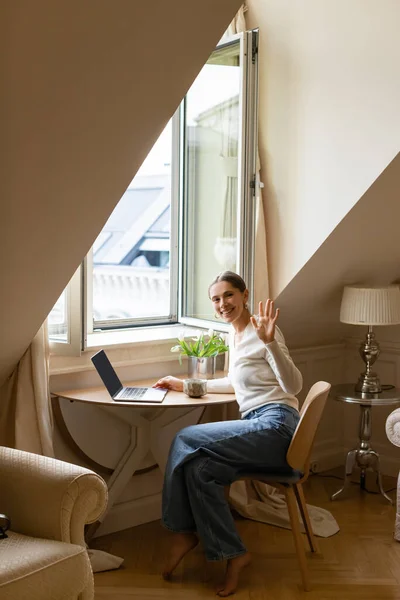 Glückliche Frau winkt Hand, während sie mit Laptop und Tonbecher am Fenster sitzt — Stockfoto