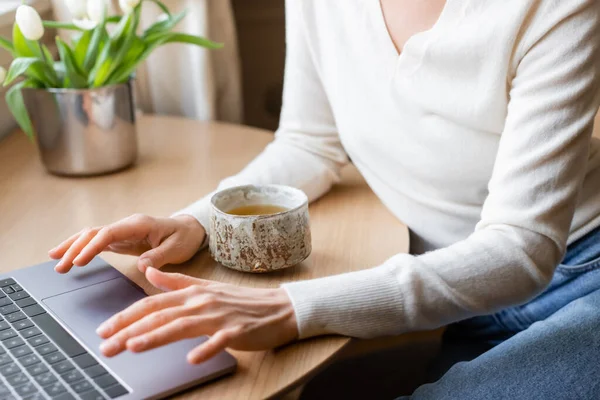 Partial view of woman typing on laptop near clay cup with tea and blurred tulips — Stock Photo