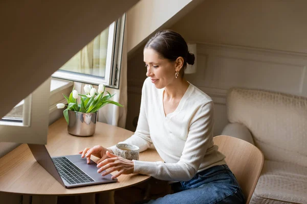 Positive woman typing on laptop near window, clay cup and fresh tulips — Stock Photo