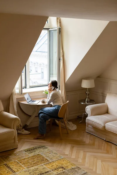 Barefoot woman talking on mobile phone near laptop and window in cozy attic room — Foto stock