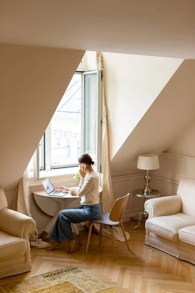 Full length of barefoot woman in jeans typing on laptop during phone conversation — Foto stock