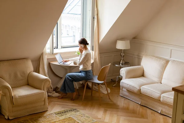 Full length view of barefoot woman talking on smartphone near laptop in attic room - foto de stock