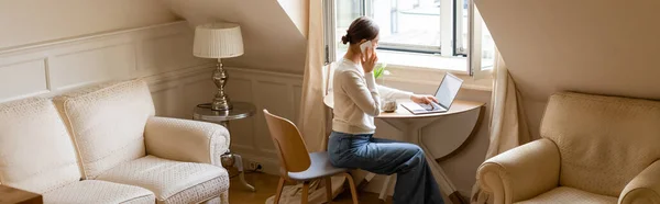 Woman in jeans talking on smartphone near laptop and window in attic room, banner — Foto stock