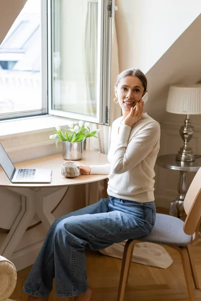 Mujer sonriendo a la cámara mientras habla en el teléfono inteligente cerca del ordenador portátil y tulipanes - foto de stock