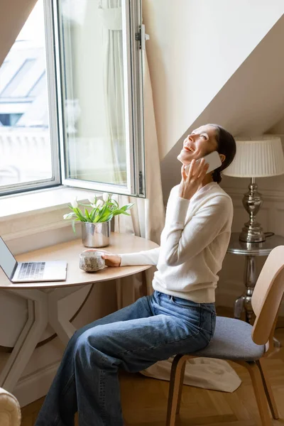 Donna eccitata ridendo durante la conversazione su smartphone vicino tazza di argilla, laptop e tulipani — Foto stock