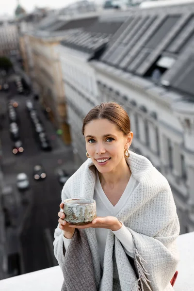 Woman covered with shawl holding clay cup and smiling at camera near rooftops on blurred background — Stockfoto