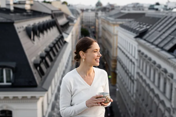 Femme souriante avec tasse d'argile regardant flous toits urbains — Photo de stock