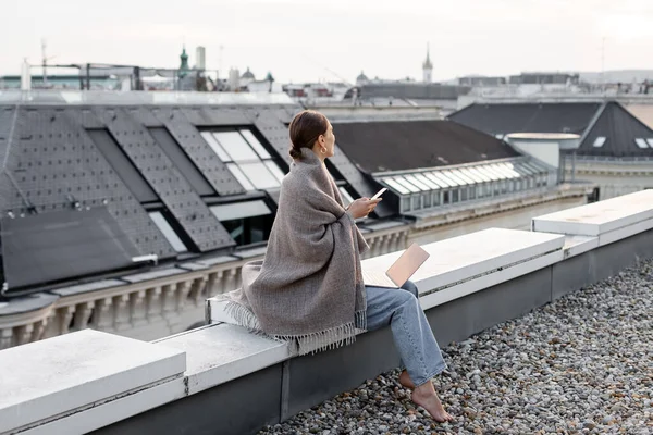 Barefoot woman covered with shawl holding smartphone near laptop on rooftop — Stock Photo